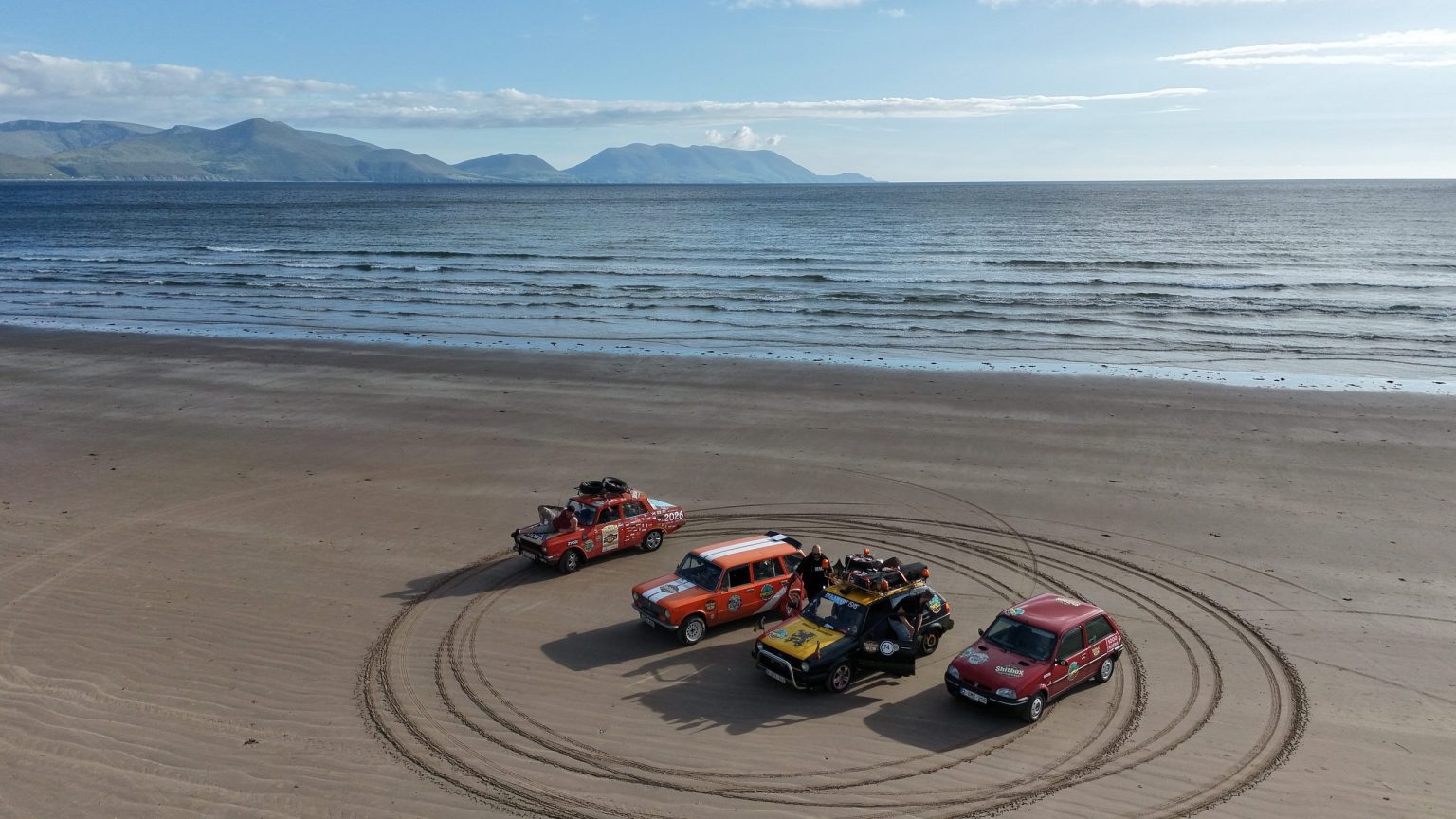 Rally cars parked in a ring of tyre tracks on a wide sandy beach