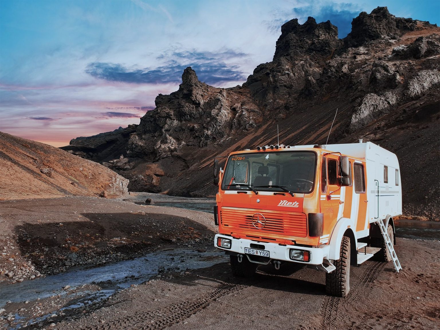 Orange and white expedition truck parked by a stream below dark lava crags