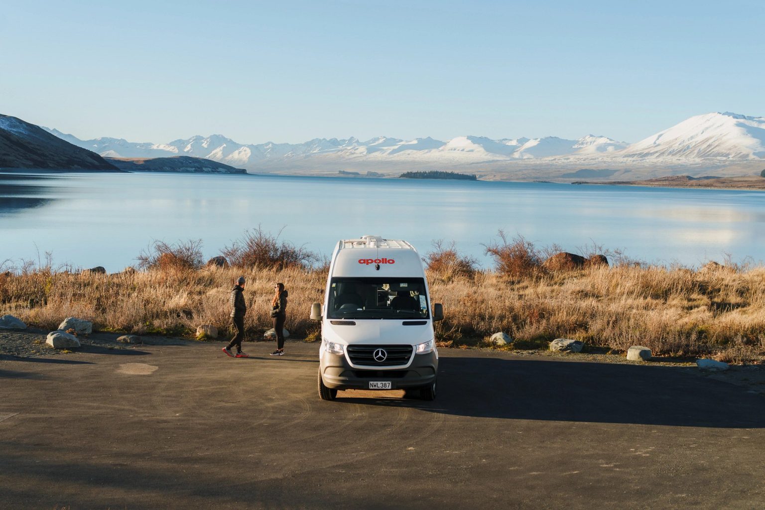Couple walking beside a campervan on a lakeshore below snowy peaks, Lake Tekapo, New Zealand's South Island