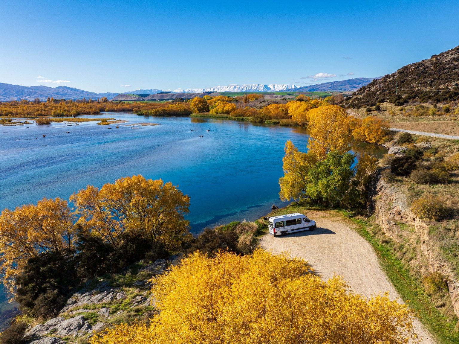 Campervan on a track beside a blue river with autumn trees