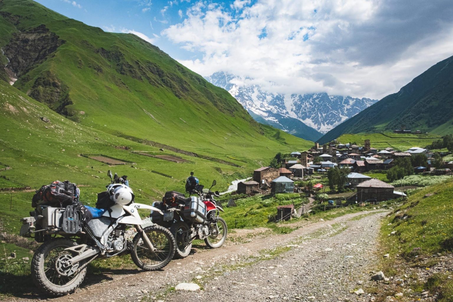 Adventure motorcycles parked above a Svaneti village with snow-capped Caucasus peaks behind