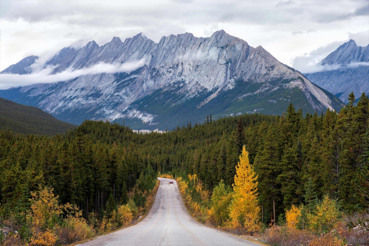 Gravel road through autumn forest toward jagged snowy peaks