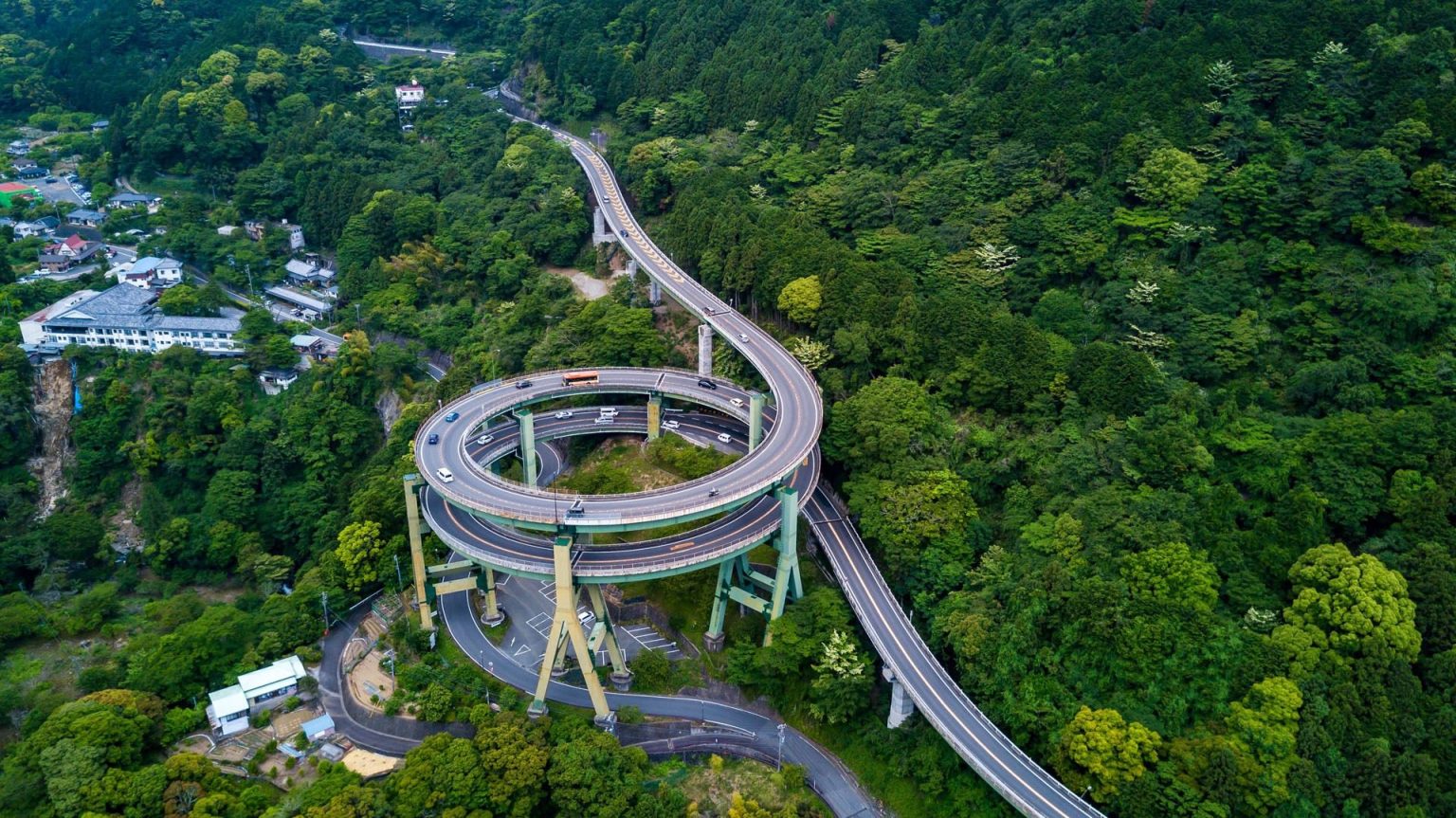 Aerial view of a looping spiral road bridge through dense green forest, Japan
