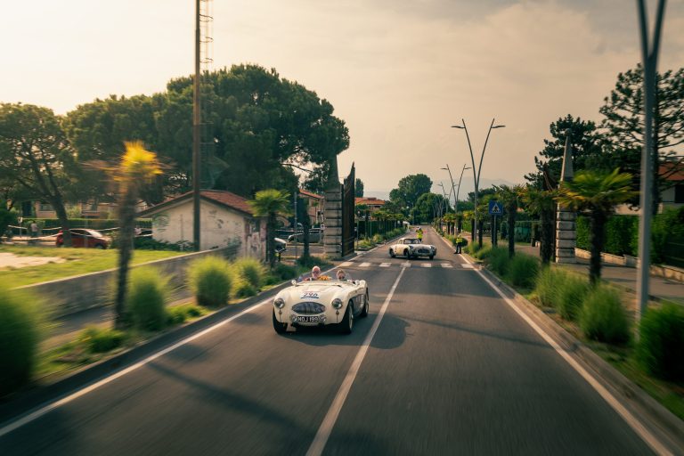 A white Austin Healey at speed on a tree-lined Italian road between control points