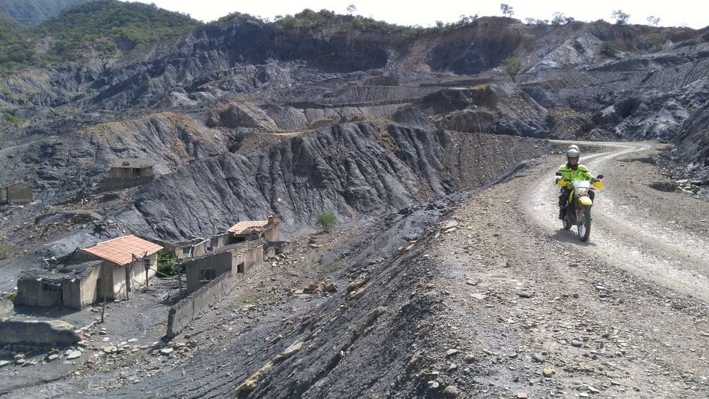 Motorcyclist on a gravel road through grey, eroded hills with a ruined village