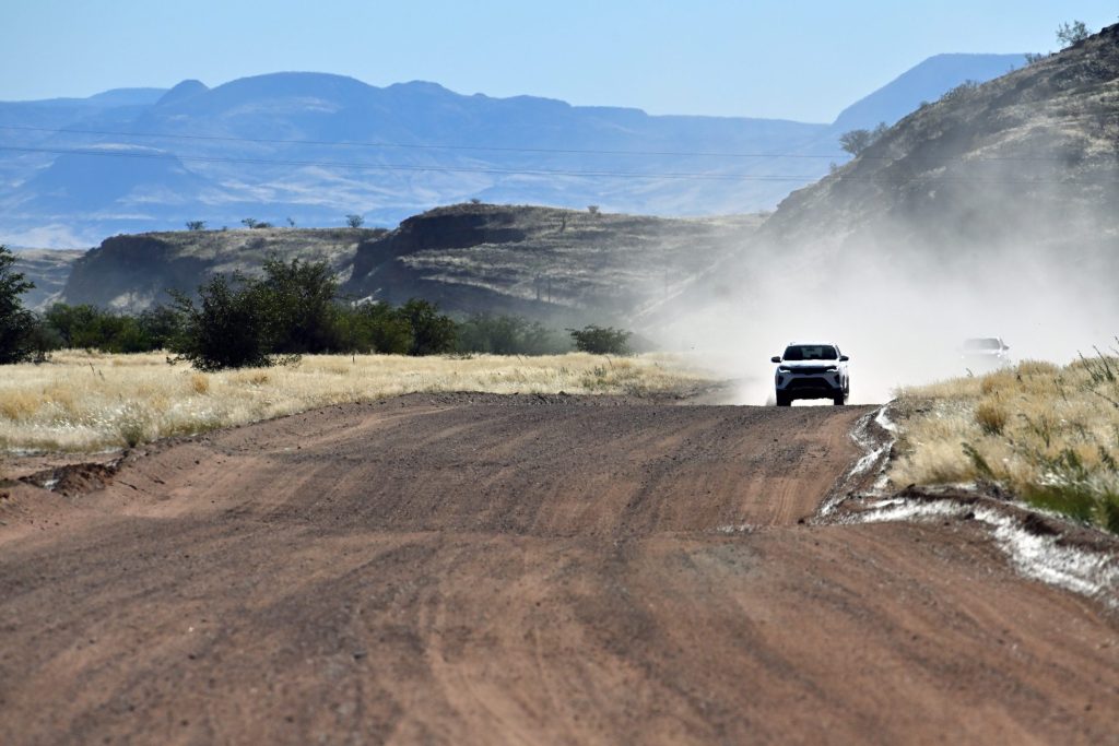 Four-wheel-drive vehicle raising dust on a wide gravel road through dry Namibian hills