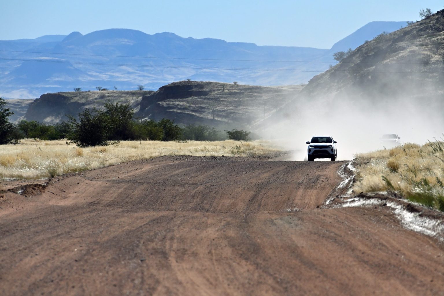 Four-wheel-drive vehicle raising dust on a wide gravel road through dry Namibian hills
