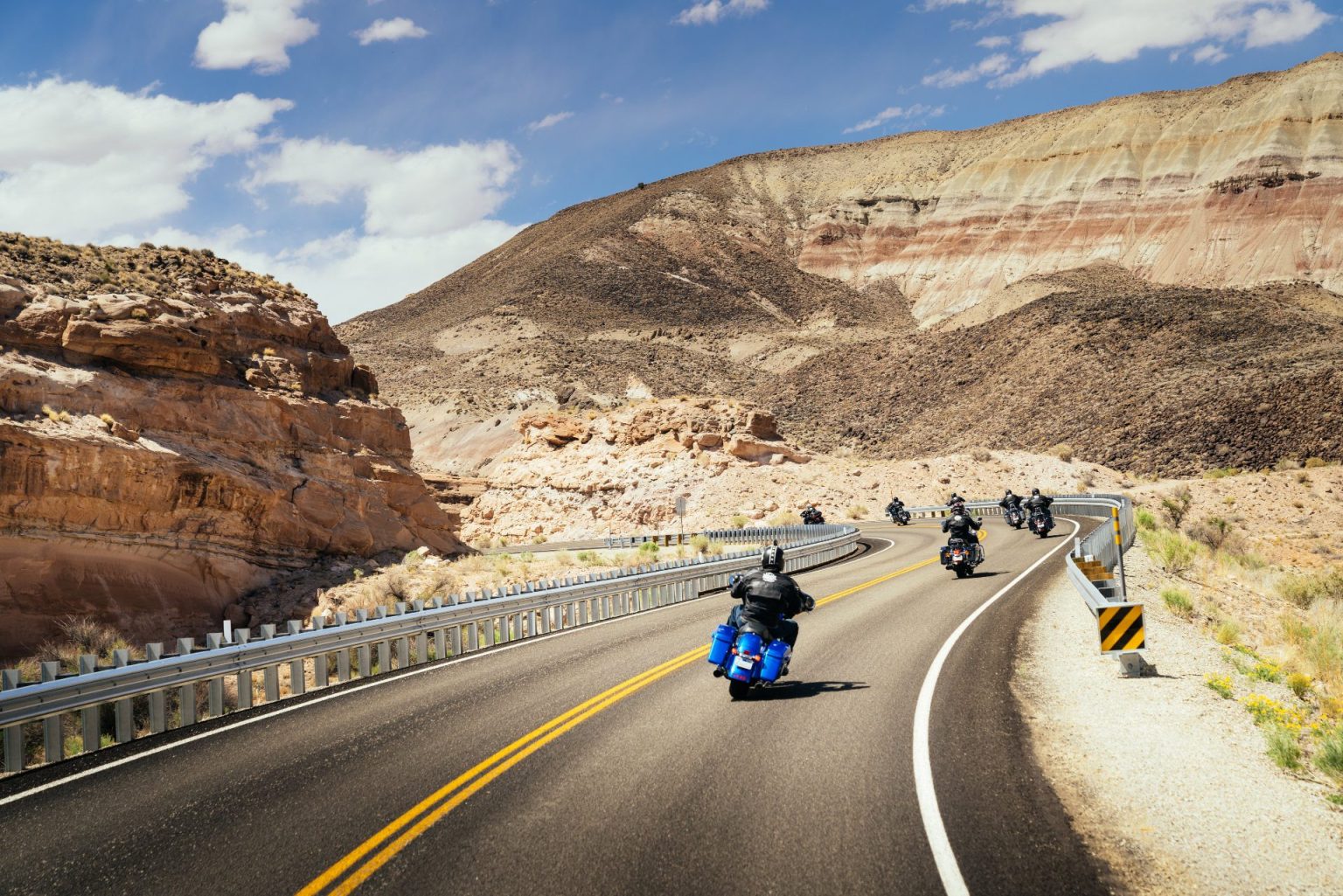 Group of motorcyclists on a curving desert road beneath striped canyon walls