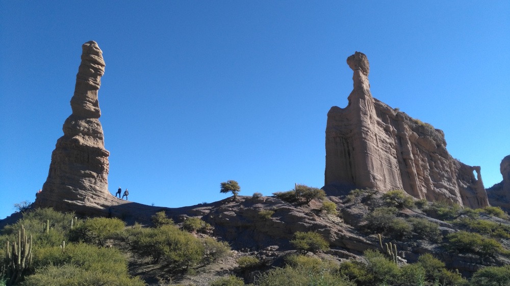 Two tall rock pinnacles above a cactus-covered hillside under a deep blue sky